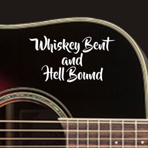 May include: A close-up of an acoustic guitar with a black and brown finish. The guitar has a soundhole and six strings. The text "Whiskey Bent and Hell Bound" is written in white on the guitar.