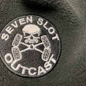 May include: A black and white embroidered patch with a skull and crossbones design. The patch reads "Seven Slot Outcast".