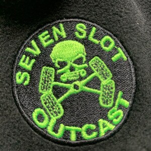 May include: A black and green embroidered patch with a skull and crossbones design. The text "SEVEN SLOT OUTCAST" is written around the skull and crossbones.