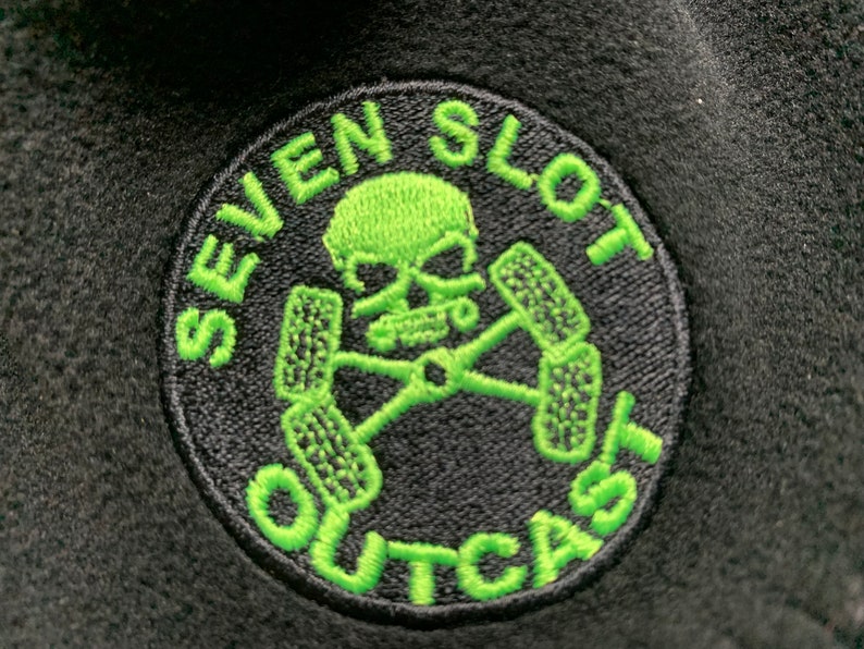 May include: A black and green embroidered patch with a skull and crossbones design. The text "SEVEN SLOT OUTCAST" is written around the skull and crossbones.