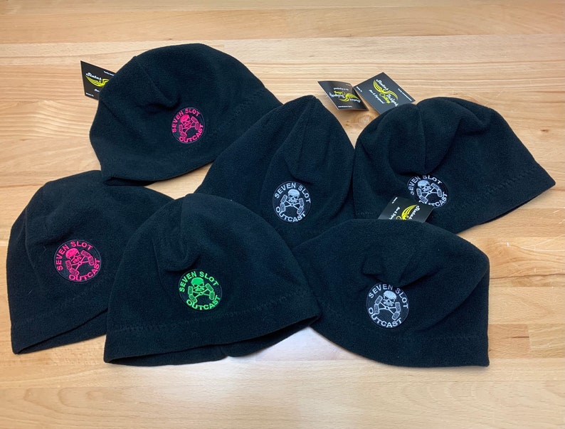 May include: Five black fleece beanies with a skull and crossbones logo that says "Seven Slot Outcast". The logos are in different colors, including pink, green, and white.