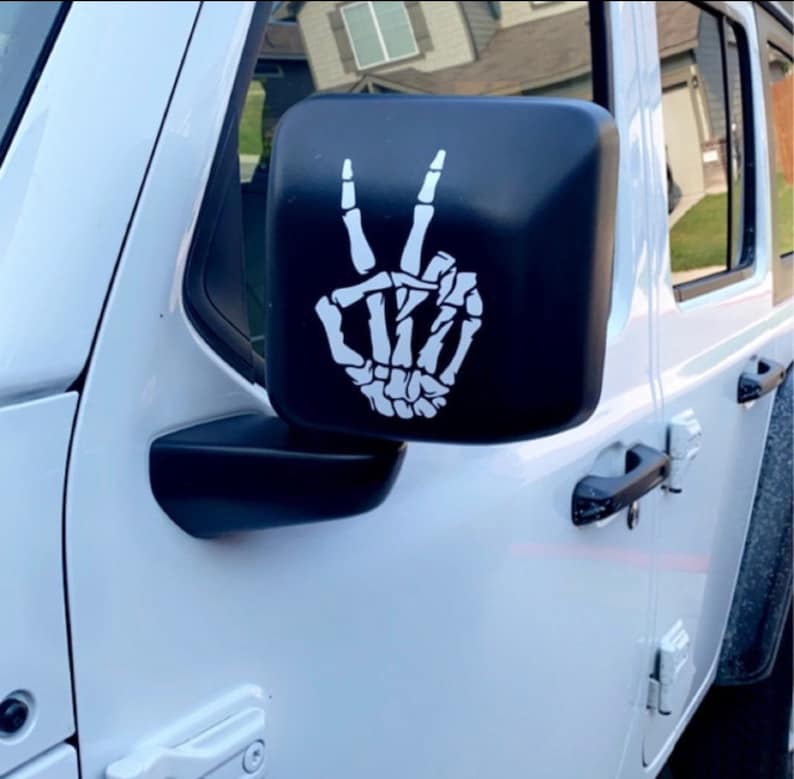 May include: A white Jeep Wrangler with a black skeleton hand peace sign sticker on the side mirror.