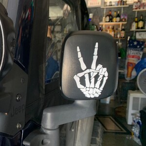 May include: A black side view mirror with a white skeleton hand making the peace sign. The mirror is attached to a black vehicle.