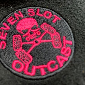 May include: A black and pink embroidered patch with the text "Seven Slot Outcast" and a skull with crossed tire tracks.