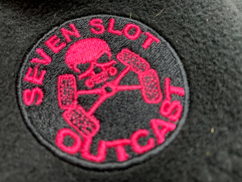 May include: A black and pink embroidered patch with the text "Seven Slot Outcast" and a skull with crossed tire tracks.