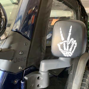 May include: A black Jeep with a skeleton hand peace sign sticker on the side mirror. The Jeep has a black top and a black door. The word "Jeep" is visible on the door.