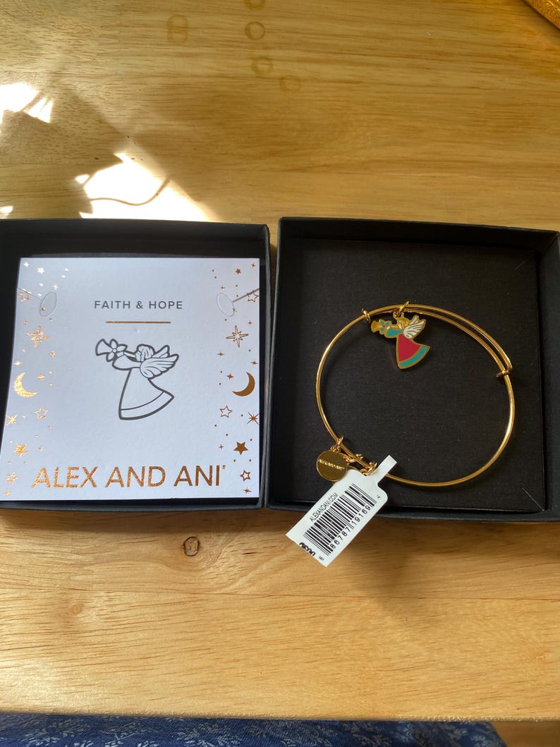 May include: A gold-coloured bangle bracelet with a charm featuring a colourful angel design. The bracelet is displayed in a black box next to a card with the text "FAITH & HOPE" and the brand name "ALEX AND ANI". A price tag is attached.
