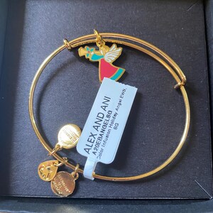 May include: Gold-coloured bangle bracelet with a colourful angel charm, a heart charm, and two round charms. The angel charm features red, teal, and gold accents. The bracelet includes a tag with the text "ALEX AND ANI".