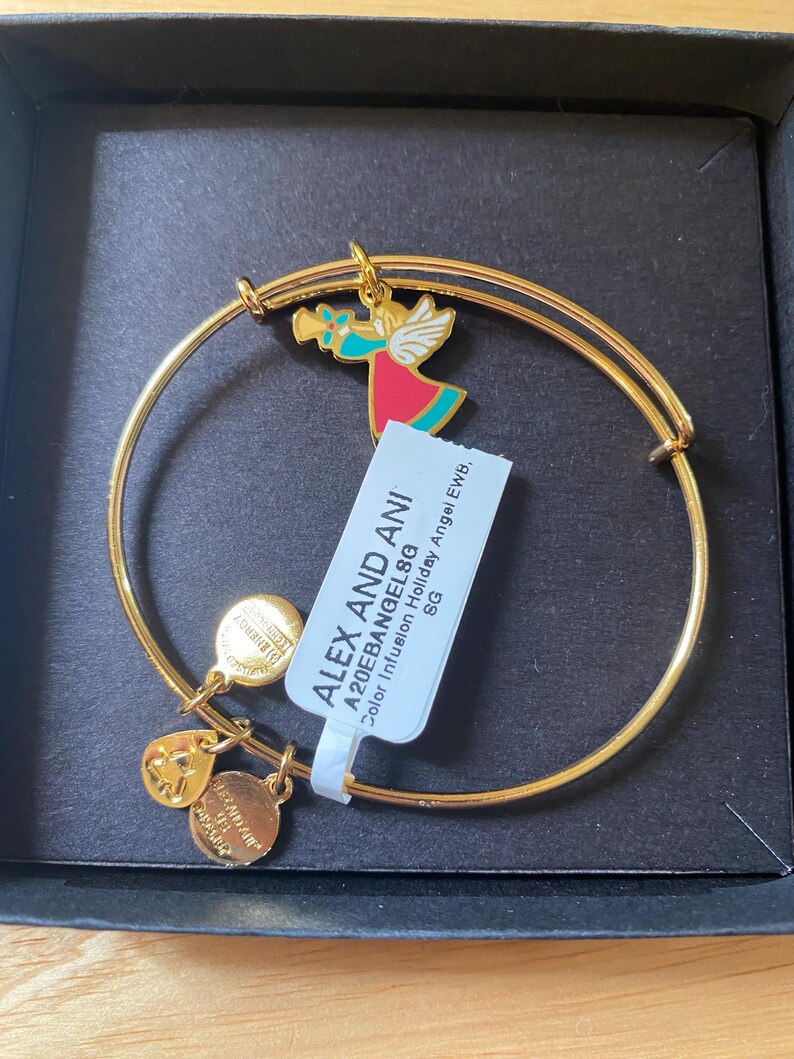 May include: Gold-coloured bangle bracelet with a colourful angel charm, a heart charm, and two round charms. The angel charm features red, teal, and gold accents. The bracelet includes a tag with the text "ALEX AND ANI".