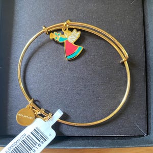 May include: Gold-tone metal bangle bracelet with a charm featuring a colourful angel playing a trumpet. The angel has white wings, a red dress, and turquoise accents. A small gold disc with the brand name is attached. The bracelet is in a black box.