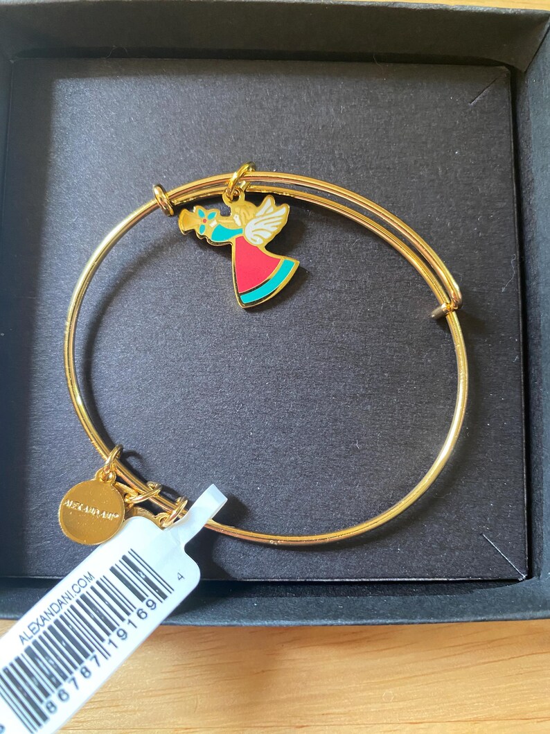 May include: Gold-tone metal bangle bracelet with a charm featuring a colourful angel playing a trumpet. The angel has white wings, a red dress, and turquoise accents. A small gold disc with the brand name is attached. The bracelet is in a black box.