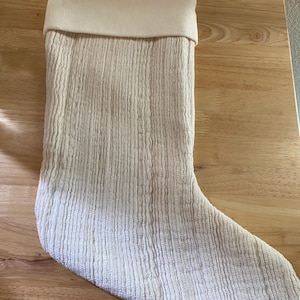May include: A cream-colored Christmas stocking featuring a textured, ribbed fabric pattern throughout the body and a smooth, solid cream-colored cuff at the top. A small white product tag is attached to the upper edge of the stocking.