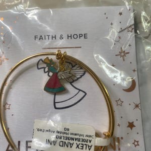 May include: A gold-tone bangle bracelet with a charm depicting a colourful angel. The angel is dressed in red and teal and holds a golden horn. The bracelet is packaged with a tag that reads "FAITH & HOPE".