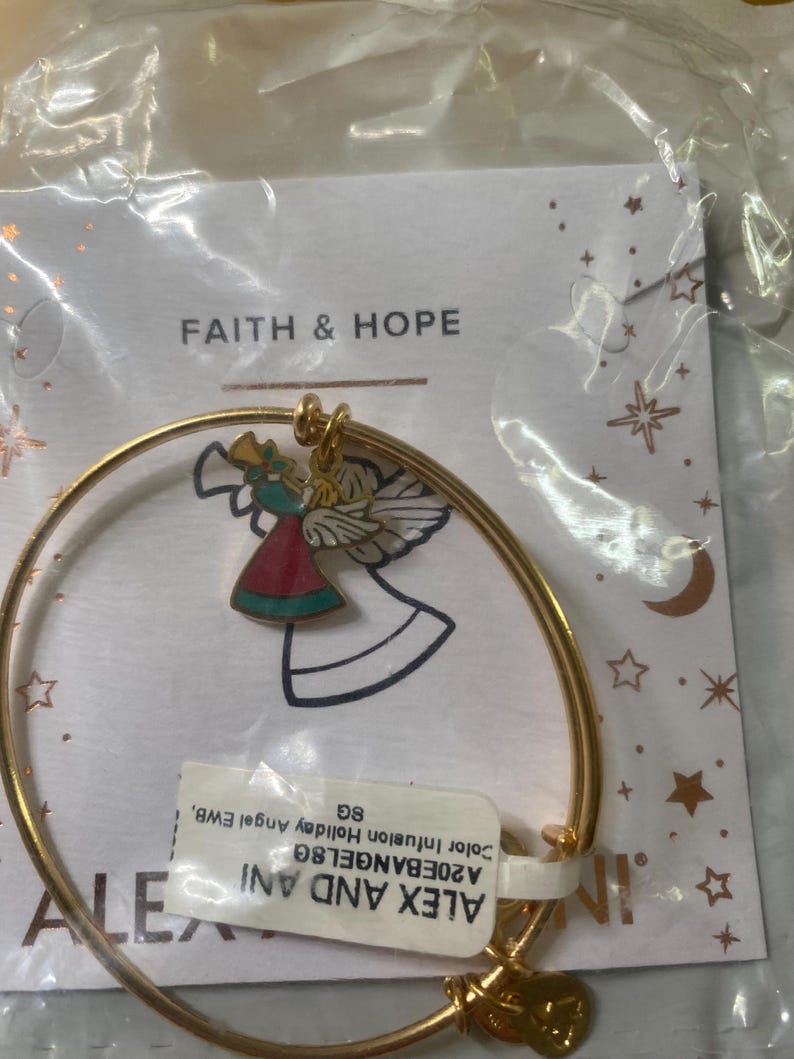 May include: A gold-tone bangle bracelet with a charm depicting a colourful angel. The angel is dressed in red and teal and holds a golden horn. The bracelet is packaged with a tag that reads "FAITH & HOPE".
