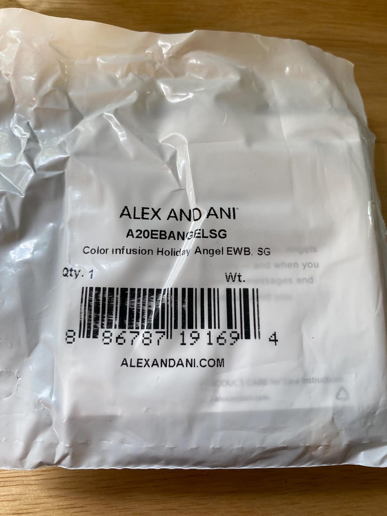 May include: A transparent plastic bag with ALEX AND ANI branding and product information. The text includes "Color infusion Holiday Angel EWB. SG" and a barcode. The bag is sealed and appears to contain a small item.