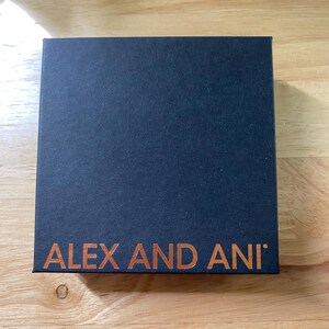 May include: A dark blue square box with the words "ALEX AND ANI" printed in a copper colour. The box is on a light brown wooden surface.