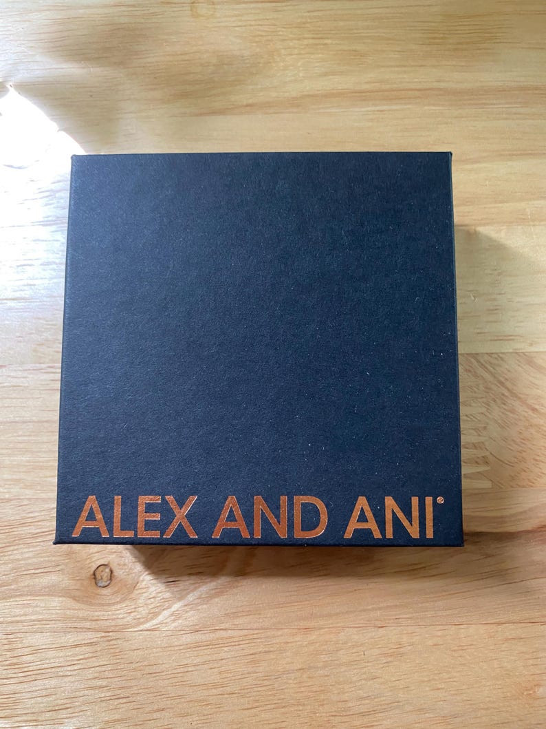 May include: A dark blue square box with the words "ALEX AND ANI" printed in a copper colour. The box is on a light brown wooden surface.