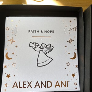 May include: A white card with the words "FAITH & HOPE" and "ALEX AND ANI" in gold lettering. An angel illustration is featured in the center. The card is decorated with gold stars and crescent moons. The card is inside a black box.
