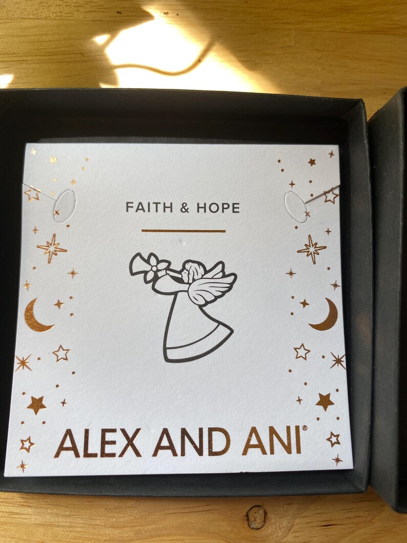 May include: A white card with the words "FAITH & HOPE" and "ALEX AND ANI" in gold lettering. An angel illustration is featured in the center. The card is decorated with gold stars and crescent moons. The card is inside a black box.