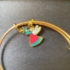 May include: Gold-coloured bangle bracelet with a charm depicting a colourful angel playing a trumpet. The angel has red, teal, and white accents. The bracelet is a simple, elegant design, suitable for everyday wear or as a gift.