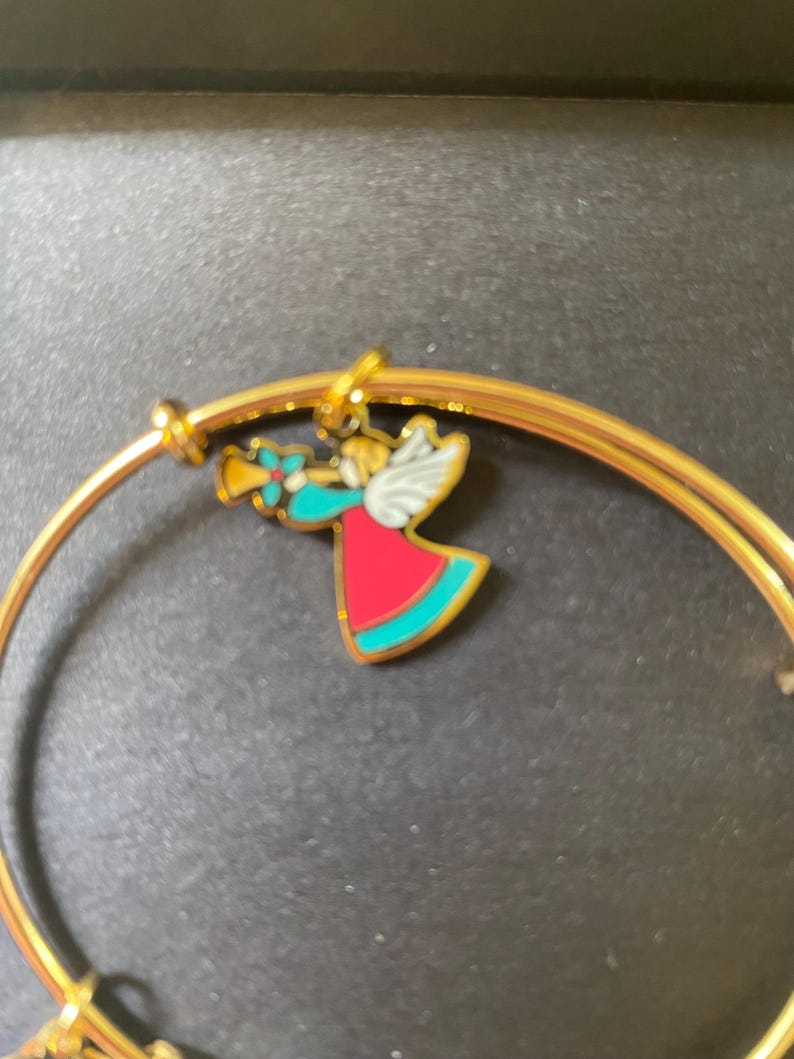 May include: Gold-coloured bangle bracelet with a charm depicting a colourful angel playing a trumpet. The angel has red, teal, and white accents. The bracelet is a simple, elegant design, suitable for everyday wear or as a gift.