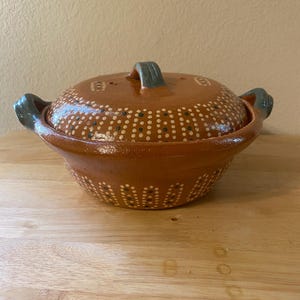 May include: A round, lidded terracotta cooking pot with a brown glaze. The pot features a series of white and green dots, with two handles and a handle on the lid. The handles and lid handle are painted a muted green.