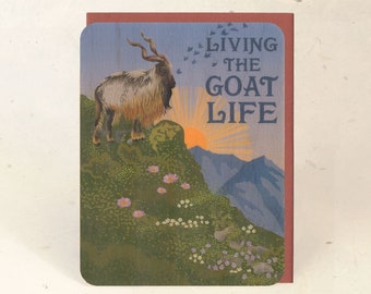 May include: Greeting card depicting a mountain goat on a hillside, with the text "LIVING THE GOAT LIFE." The illustration has a vintage style, with a sunset, mountains, and birds in the background. The card has a red border.