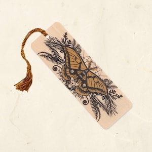 May include: A rectangular wooden bookmark featuring a black ink illustration of a large moth surrounded by fern leaves and geometric lines. The bookmark includes a hole at the top with a metallic gold cord tassel attached to the end.