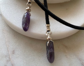 May include: A black suede cord necklace with two amethyst stone pendants. The stones are wrapped in silver wire and have a pointed shape.