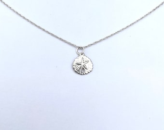 May include: A delicate silver necklace featuring a small, detailed sand dollar pendant. The pendant is round with a star design in the center and intricate patterns. The chain is thin and silver, creating a minimalist and elegant look.