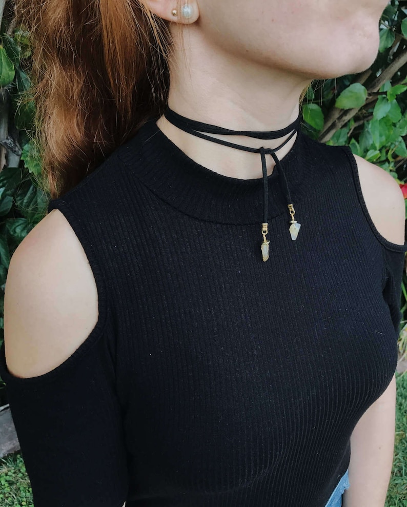 May include: A black choker necklace with two small, light-colored stones hanging from it. The necklace is worn by a person with red hair.