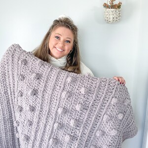 May include: A soft, light purple crocheted blanket with a textured bobble stitch pattern. The blanket is held by a person in front of a white wall.