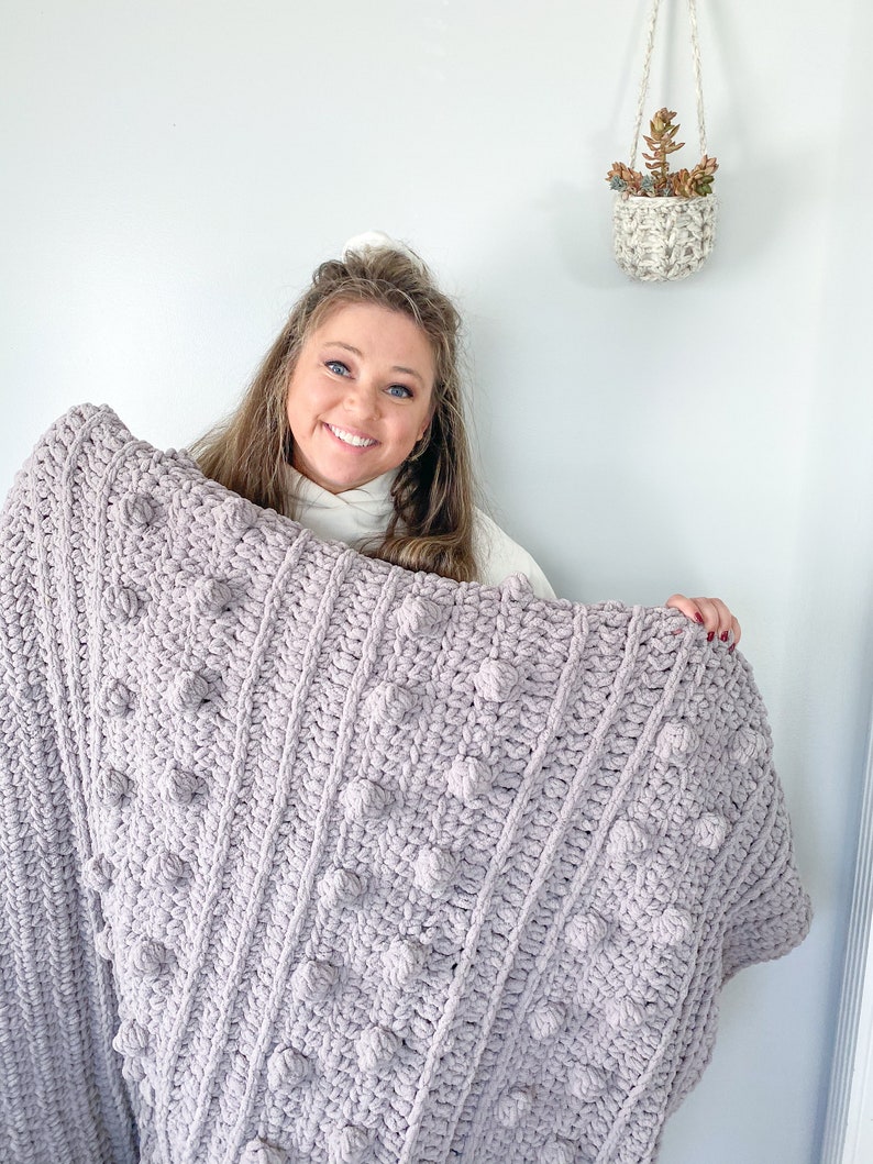 May include: A soft, light purple crocheted blanket with a textured bobble stitch pattern. The blanket is held by a person in front of a white wall.