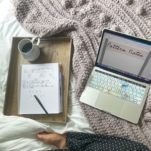 May include: A laptop computer with a white keyboard and a screen displaying the text "Pattern Notes". A notebook with handwritten notes about rows and repeats is on a wooden tray next to a cup of coffee.