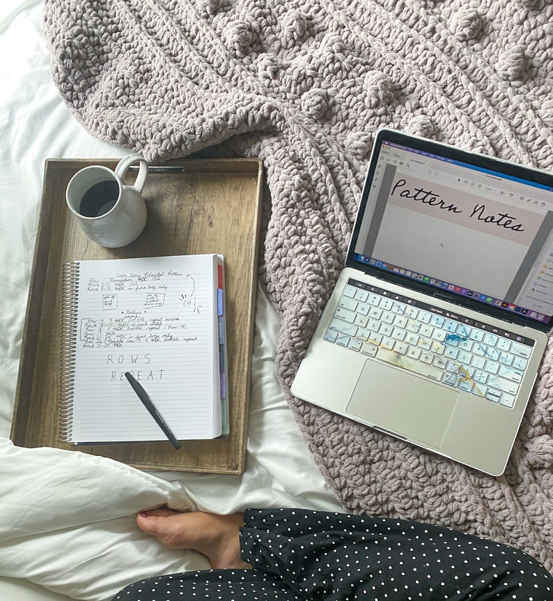 May include: A laptop computer with a white keyboard and a screen displaying the text "Pattern Notes". A notebook with handwritten notes about rows and repeats is on a wooden tray next to a cup of coffee.