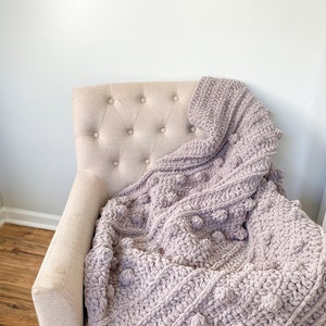 May include: A soft, plush, light purple throw blanket with a textured, bobble stitch pattern. The blanket is draped over a beige armchair.