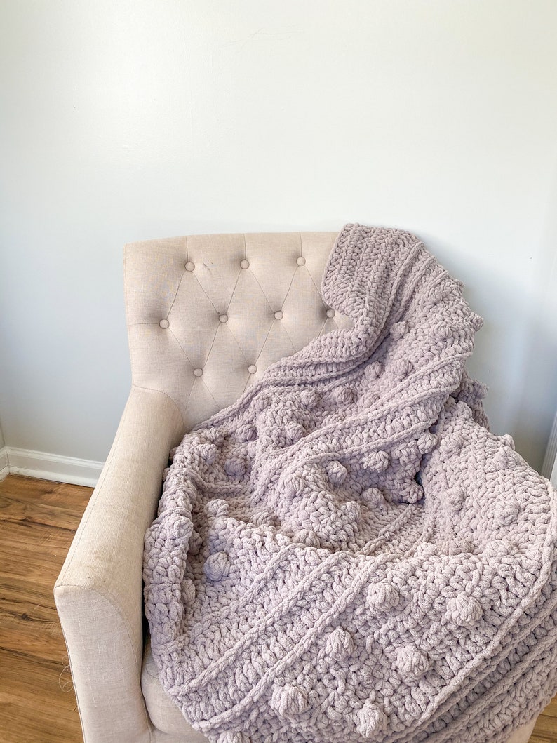May include: A soft, plush, light purple throw blanket with a textured, bobble stitch pattern. The blanket is draped over a beige armchair.