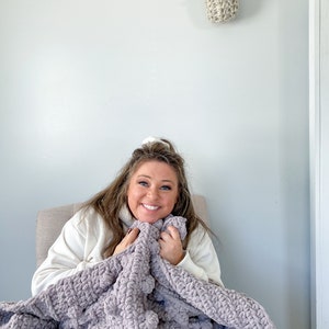 May include: A person sitting in a chair, wrapped in a soft, plush, gray blanket. The blanket has a textured, chunky knit design.