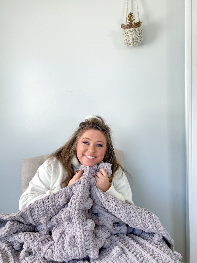 May include: A person sitting in a chair, wrapped in a soft, plush, gray blanket. The blanket has a textured, chunky knit design.