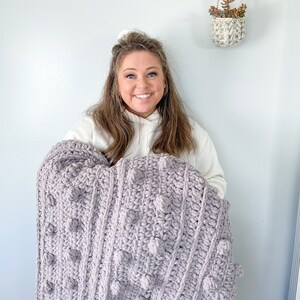 May include: A woman holds a large, soft, purple crocheted blanket with a textured, bobble stitch pattern.