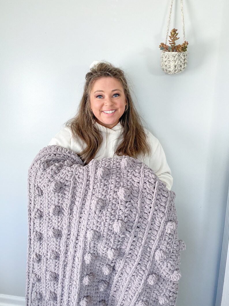 May include: A woman holds a large, soft, purple crocheted blanket with a textured, bobble stitch pattern.