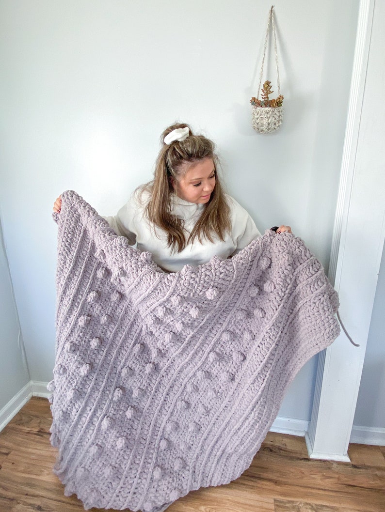 May include: A woman holds up a large, crocheted blanket with a textured, bobble stitch pattern. The blanket is a light purple color.