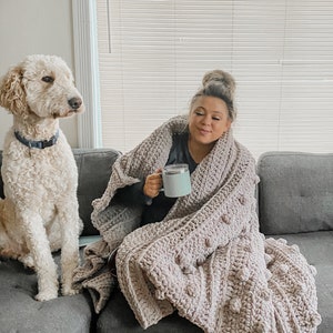 May include: A woman is sitting on a couch with a dog. She is wrapped in a large, light pink crocheted blanket with large, round, textured stitches. She is holding a light blue mug in her hand.