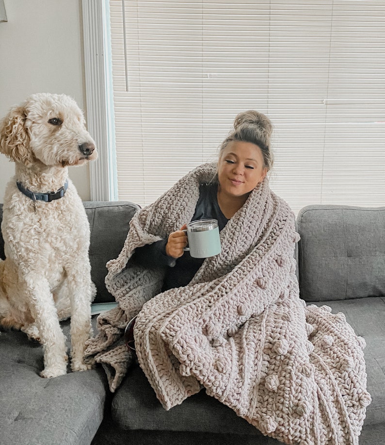May include: A woman is sitting on a couch with a dog. She is wrapped in a large, light pink crocheted blanket with large, round, textured stitches. She is holding a light blue mug in her hand.