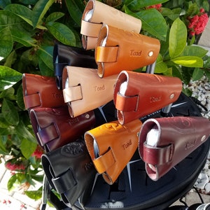 May include: A close-up of a set of nine leather golf club headcovers in various shades of brown, tan, and burgundy. Each headcover features the word "Toad" embossed on the side. The headcovers are arranged in a stack on a black golf bag.