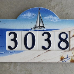 May include: A white ceramic house number sign with a blue and white seascape scene. The number 3038 is painted in blue on the sign. The sign has a white border and a scalloped top.