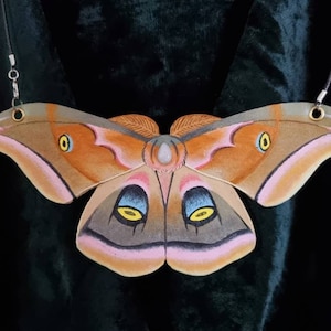 Pode incluir: Um colar de mariposa pintado em marrom e rosa com uma pedra branca no centro. A mariposa tem olhos amarelos e está pendurada em uma corrente prateada.