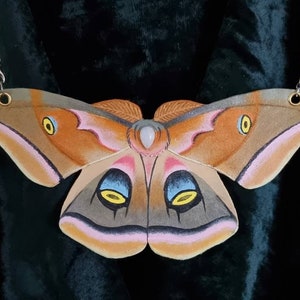 Pode incluir: Um colar de madeira pintado de marrom e rosa em forma de borboleta com uma pedra branca no centro. A borboleta tem olhos amarelos e azuis.
