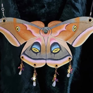 Pode incluir: Um colar grande em forma de mariposa pintada com detalhes intrincados e pingentes pendurados. A mariposa é pintada em tons de marrom, rosa e cinza com detalhes amarelos. Os pingentes são feitos de contas roxas e douradas e apresentam pequenas estrelas.