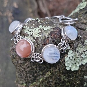 May include: A silver bracelet featuring a series of round, bezel-set gemstones. The bracelet alternates between peach and iridescent blue-white stones, connected by small silver links. The clasp is a simple toggle style.